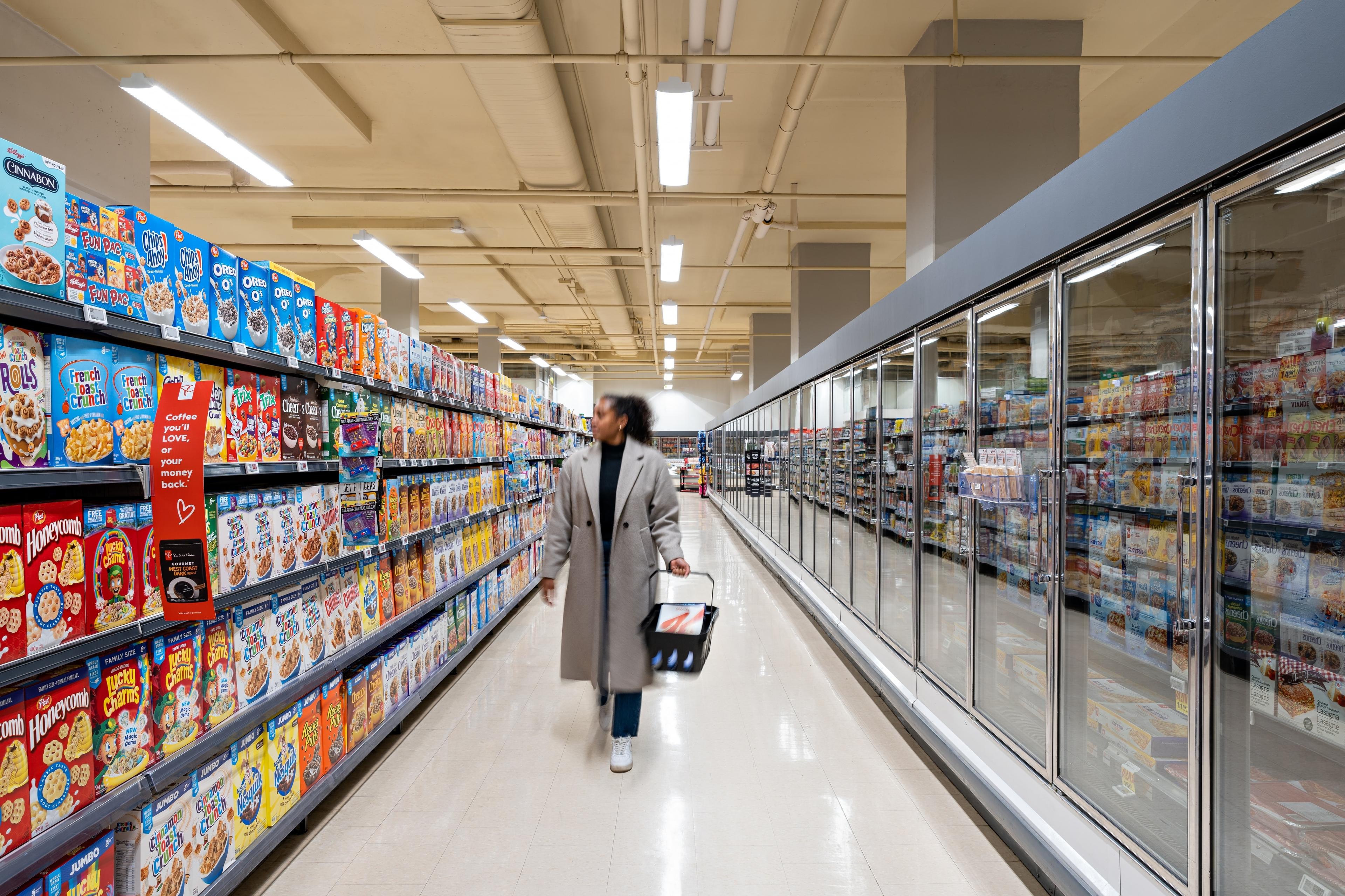 A woman is walking down a grocery store aisle looking at cereals. 
