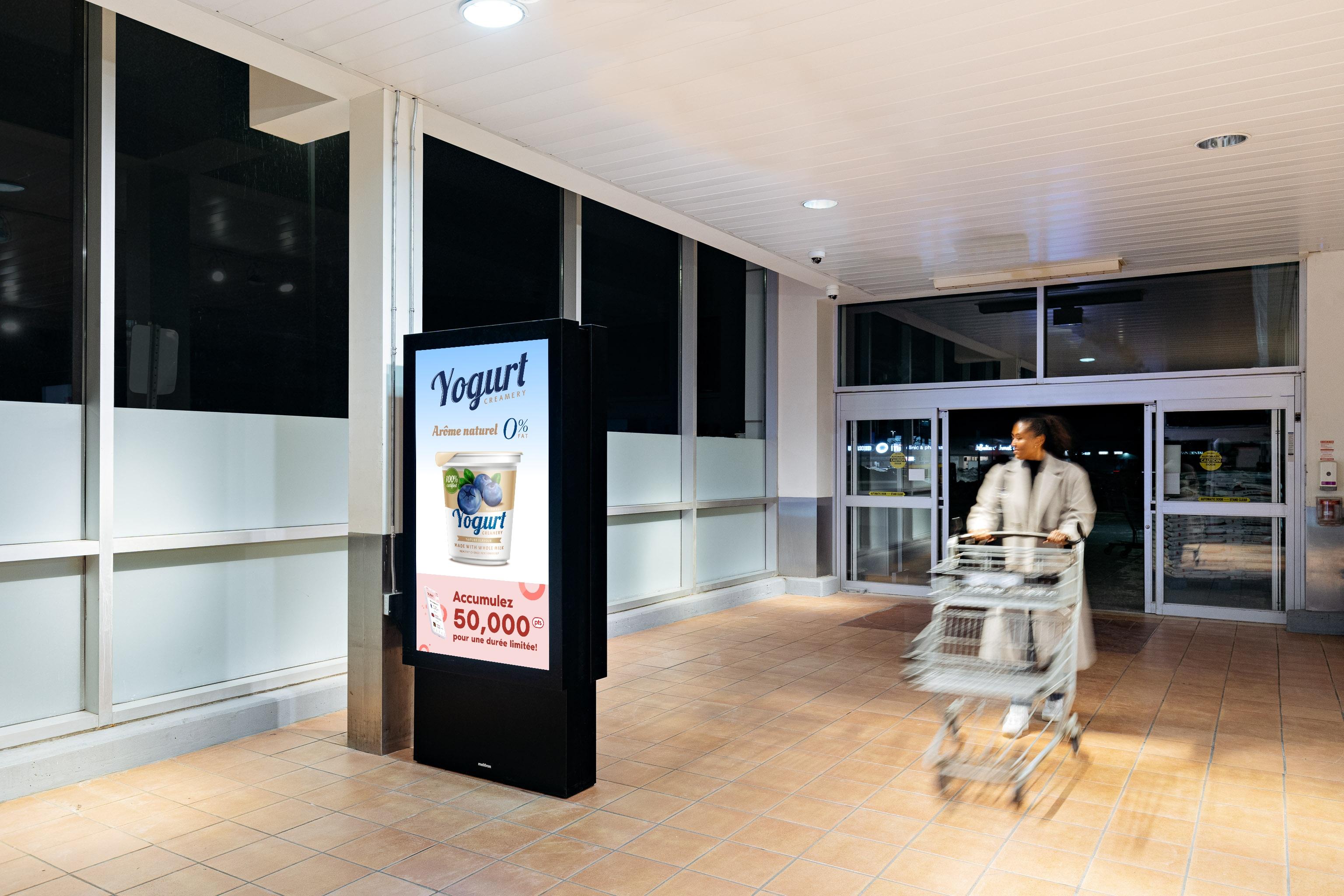 Une femme passe devant un écran à l'entrée principale d'une épicerie avec une publicité pour un yaourt dessus.
